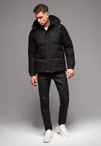 Svart vadderad jacka med huva, som har ett subtilt mönster och guldtonade detaljer. Kombinerad med svarta byxor och vita sneakers.