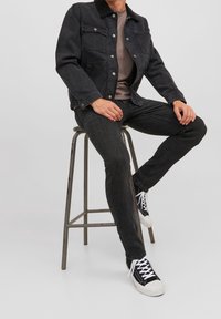 Svart denimjacka med två bröstfickor, bärs över en grå skjorta, i kombination med mörka jeans och svarta sneakers med vita sulor, sittande på en pall.