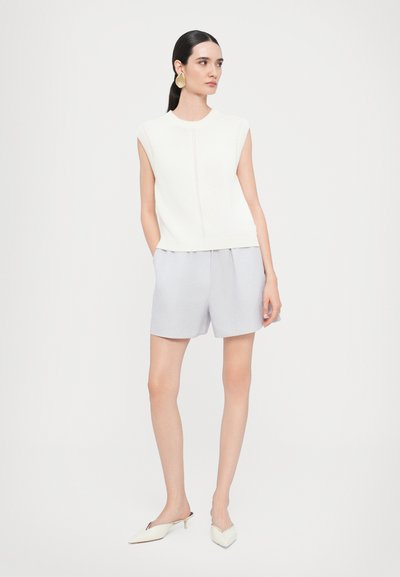 Top senza maniche color beige chiaro in maglia con scollatura rotonda, abbinato a pantaloni corti grigi chiari. Calzature sono mule bianchi a punta. Design minimalista.