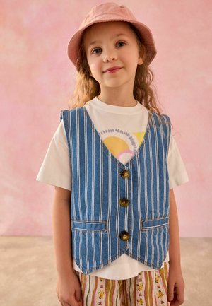 Niña joven con un sombrero rosa, camiseta blanca con diseño, chaleco azul a rayas y pantalones con rayas y flores sobre un fondo rosa.