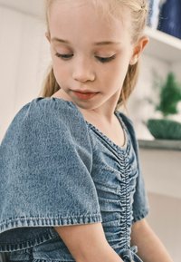 Blå denimtop med korte pufærmer, der har et samlet design og en bindedetalje ved taljen. Glat tekstur og afslappet stil.