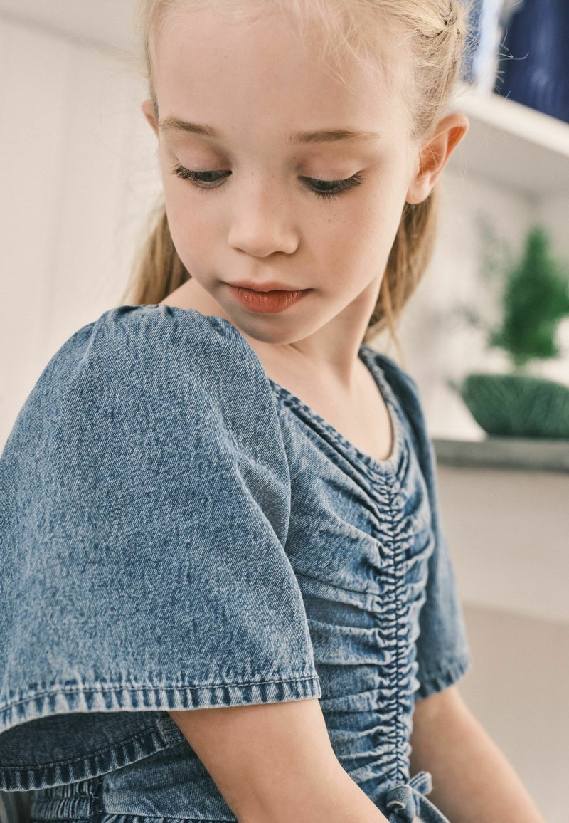 Blå denimtop med korte pufærmer, der har et samlet design og en bindedetalje ved taljen. Glat tekstur og afslappet stil.