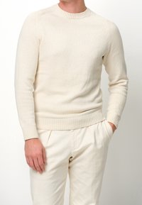 Maglione lavorato a maglia color crema con scollo a girocollo e polsini a costine. Orlo arrotondato e vestibilità aderente. Abbinato a pantaloni color crema.