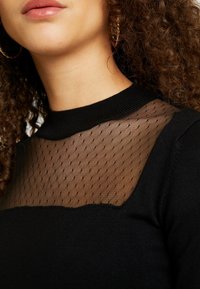 Nainen, joka pukeutunut mustaan yläosaan, jossa on läpikuultava leikkaus ja pilkullinen kuvio kauluksen lähellä, kiharat hiukset ja näkyvät kultaiset renkorenkorvikset.