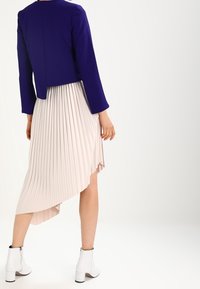 Fialová krátká bunda s dlouhými rukávy, doplněná béžovou plisovanou sukní s asymetrickým lemem. Bílé kotníkové boty.