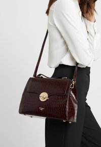 Burgundy faux krokodilläderhandväska med guldfärgad hårdvara, strukturerad form, topphandtag och avtagbar axelrem.