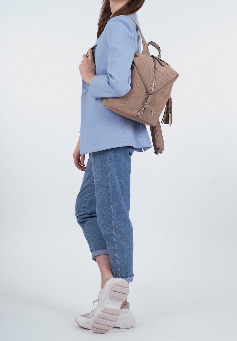 Femme portant un blazer bleu clair et un jean, portant un sac à dos beige texturé, debout avec un pied légèrement levé sur un fond uni.