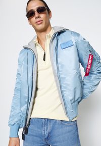 Jonge man met donkere zonnebril, lichtblauwe bomberjas met rood label op de mouw, crèmekleurig ritsshirt en blauwe spijkerbroek tegen een witte achtergrond.