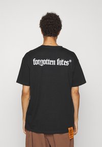 Sort bomulds T-shirt med hvid tekst på ryggen, "forgotten ftes," og et orange label på siden. Casual pasform, korte ærmer.
