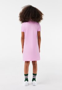 Růžové krátké šaty s rovný střih, hladkou texturou a jednoduchým designem, doplněné zelenobílými pruhovanými ponožkami a bílými teniskami.