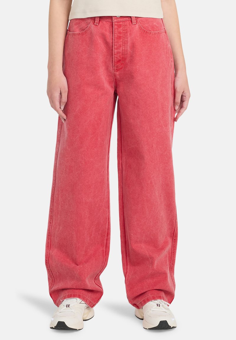 Osoba nosząca luźne, czerwone spodnie z wysokim stanem i przednimi kieszeniami oraz białe trampki, stojąca na białym tle.