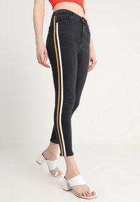 Svarta denim skinny jeans med vita och gula sidostripes. Har en hög midja och en smal passform. Bärs med vita blockklacksandaler.