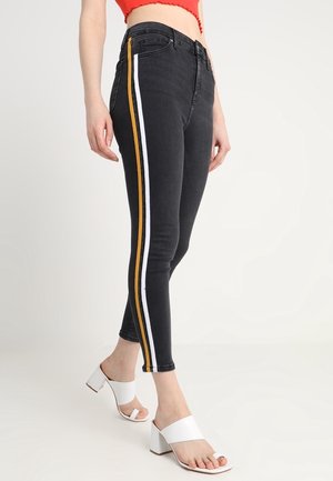 Jeans ajustados de mezclilla negra con rayas laterales blancas y amarillas. Presenta una cintura alta y un corte slim. Combinados con sandalias de tacón bloque blancas.