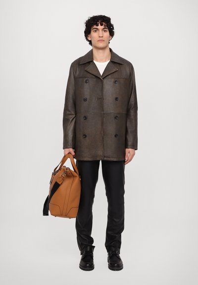 Abrigo de cuero marrón de doble botonadura con grandes botones negros, llevado sobre una camisa blanca. Pantalones negros ajustados y llevando un bolso marrón-naranja.