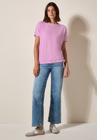 Růžové pruhované tričko s volným střihem a detailním uzlem na boku, spárované s širokými světle modrými džíny a šedými teniskami. Jednoduchý design a ležérní styl.