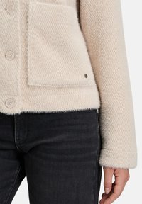 Crème-kleurige textuurjacket met een zachte, vlezige afwerking, voorzien van vier knopen en een patchzak. Gecombineerd met donkere jeans.