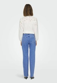 Biała koszula z długim rękawem z wyciętym kwiatowym wzorem na ramionach, zestawiona z niebieskimi jeansami o wysokim stanie, dopasowanymi, lekko wyblakłymi, o długości do kostek.
