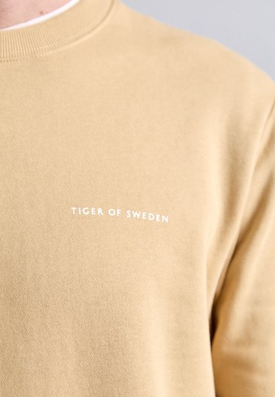 Világos bézs pamut pulóver, kerek nyakú és bordás mandzsettákkal. A mellkason fehér színnel szerepel a "TIGER OF SWEDEN" felirat.