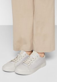 Baskets gris clair avec une finition lisse et mate ; design à lacets ; accents légers contrastants ; portées avec un pantalon beige à jambes larges.