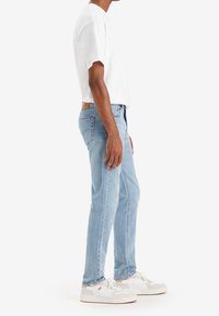 Vaqueros de mezclilla de color azul claro, diseño de pierna recta, combinados con una camiseta blanca y zapatillas blancas. Etiqueta de la cintura visible en los vaqueros.