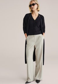 Cardigan lungo nero sopra una camicia nera con bottoni, abbinato a pantaloni largi di colore grigio chiaro e tacchi neri appuntiti. Texture lisce, design moderno.