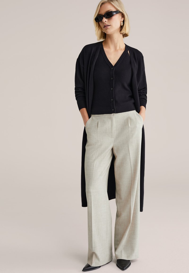 Cardigan lungo nero sopra una camicia nera con bottoni, abbinato a pantaloni largi di colore grigio chiaro e tacchi neri appuntiti. Texture lisce, design moderno.