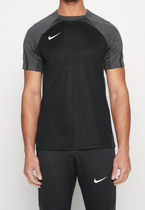 Homme portant une tenue de sport noire Nike avec un t-shirt à manches courtes aux manches grises à motifs et un pantalon noir assorti avec des logos Nike blancs.