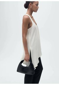 Cream-colored, sleeveless top with a V-neck and side slits, paired with black pants. Holds a textured, black woven handbag.