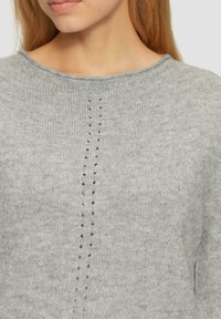 Pull en tricot gris clair avec un col rond et des trous verticaux le long du centre. Texture douce avec bordures côtelées au niveau du col.