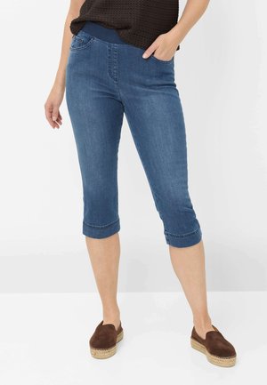 Frau trägt blaue kurze Jeanshosen mit umgeschlagenen Säumen, dunkelbraune Slip-On-Espadrilles und ein dunkles Strickoberteil, steht mit einer Hand in der Tasche.