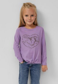 Lavender langemouw shirt met ronde halslijn, voorzien van een paardenafbeelding en tekst in een donkerdere kleur, met een knoop aan de onderkant. Gecombineerd met blauwe jeans.