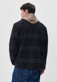 Chaqueta de cuadros en tonos navy con un patrón de rayas negras y capucha beige. Presenta un corte relajado y una tela suave, adecuada para un uso casual.