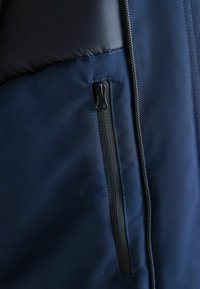 Navy blue jacket featuring a smooth fabric, padded upper section, and a zippered pocket with a black zipper on the left side.