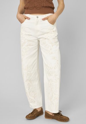 Femme portant un pantalon blanc avec broderie florale beige, un top marron texturé sans manches, et des mocassins marron, mains dans les poches.