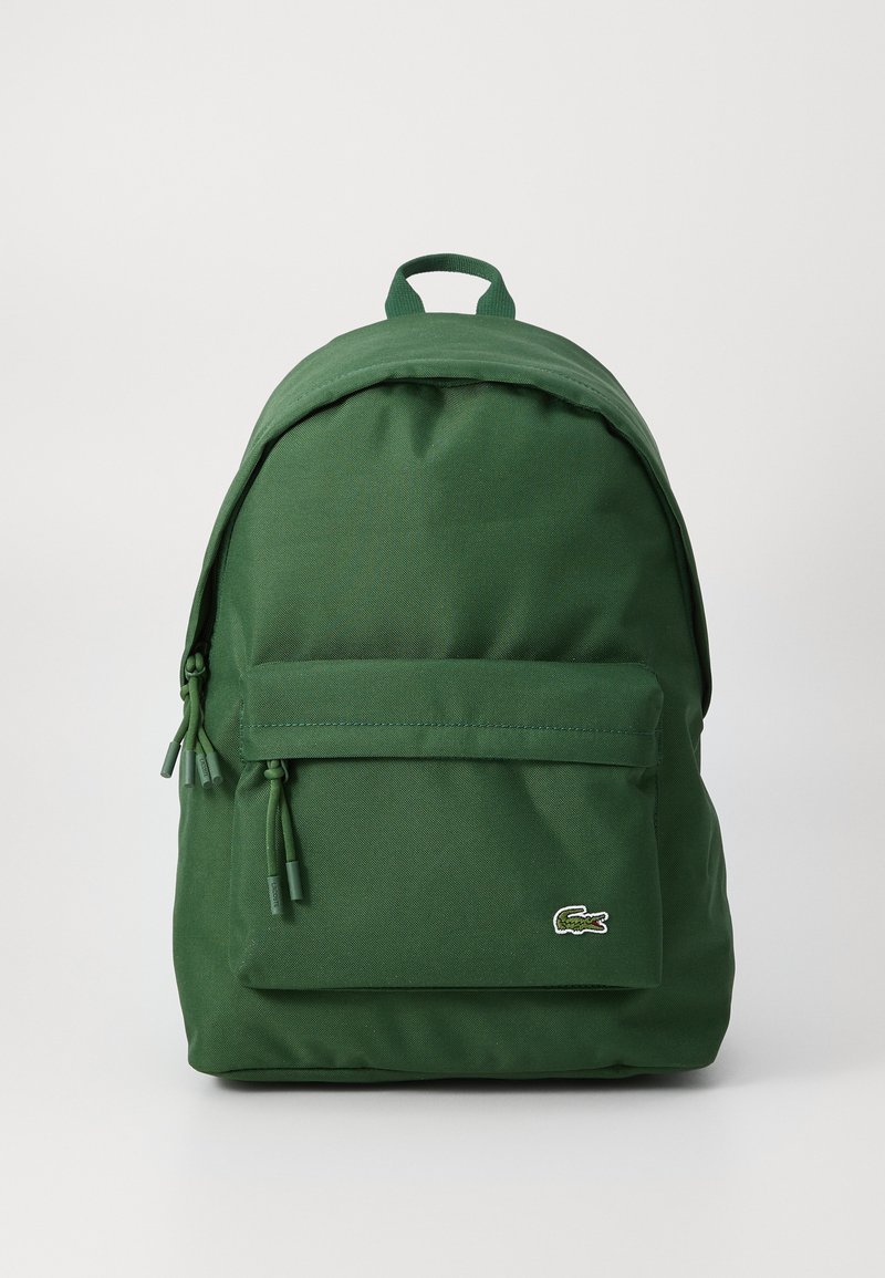 Rucsac din țesătură verde, cu formă rotunjită, buzunar frontal cu fermoar și mâner superior. Prezintă accente cu snur și un logo mic. Design simplu.