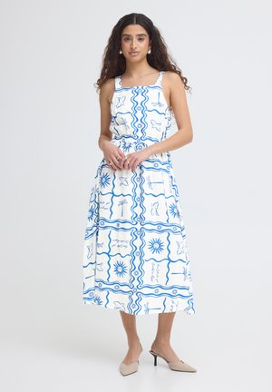 Vrouw met lang krullend haar draagt een witte mouwloze midi-jurk met een patroon van blauwe zon, vogel en golven, beige slingback hakken en pareloorbellen.
