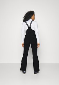 Schwarze Latzhose mit verstellbaren Trägern, die ein Logodesign aufweist, weiter Schnitt und Reißverschlusstaschen. Getragen über einem langärmeligen weißen Oberteil.