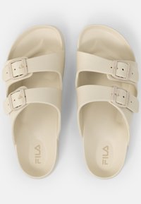 Sandales beiges avec deux larges brides maintenues par des boucles réglables, présentant une texture lisse et un léger soutien de la voûte plantaire. Logo "Fila" visible.