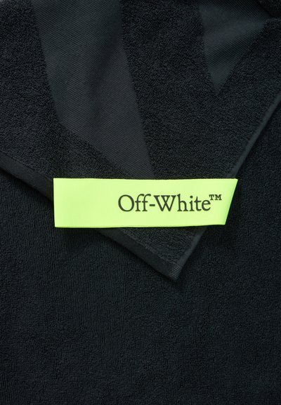 Μαύρο υλικό με υφή και διπλωμένη γωνία που δείχνει μια φωτεινή κίτρινη-πράσινη ετικέτα με την ένδειξη "Off-White™" σε μαύρο κείμενο.