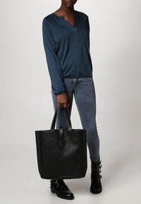 Schwarze Leder-Umhängetasche in quadratischer Form, mit robusten Griffen und einem schlichten Äußeren. Das Modell trägt ein blaues Langarmshirt und Jeans.