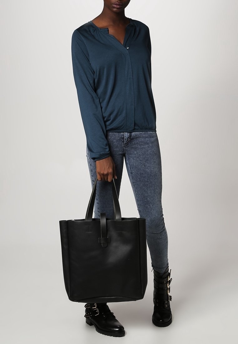 Schwarze Leder-Umhängetasche in quadratischer Form, mit robusten Griffen und einem schlichten Äußeren. Das Modell trägt ein blaues Langarmshirt und Jeans.