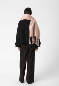 Chaqueta negra corta con un corte holgado y detalles en los puños, combinada con una bufanda larga, suave y de flecos en un delicado tono rosa claro, sobre un fondo liso.