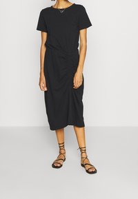 Robe t-shirt noire avec une taille cintrée, des fronces à l'avant, des manches courtes et une longueur sous le genou. Associée à des sandales noires à lanières.