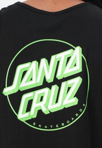 Mustas tank top, millel on suur ja julge roheline graafika, kus on kirjas "SANTA CRUZ" stiliseeritud fontide ja "SKATEBOARDS" väiksema tekstina all.