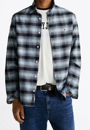 Mand iført åben lyseblå og sort ternet skjorte over hvidt grafisk t-shirt, blå jeans og sort bælte, hænder delvist i lommerne.