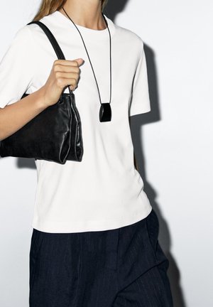 Personne portant une chemise blanche à manches courtes et un pantalon foncé, tenant un sac à main noir et portant un collier pendentif noir.
