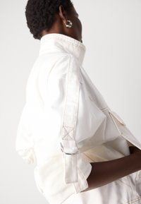 White denim jacket featuring a high collar, adjustable cuffs with metal hardware, and contrasting stitching. Smooth texture with pocket details.