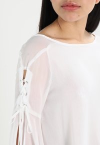 Camicia bianca, in tessuto leggero, presenta un design con lacci e grommet metallici alle aperture delle maniche, texture a contrasto e vestibilità ampia.