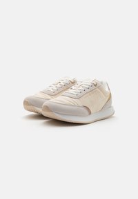 Beige sportcipők, amelyek velúrból és nylonból készültek, texturált nyelvvel, fehér cipőfűzőkkel, puha szürke részletekkel és gumitalppal.