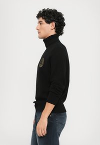 Črni pulover s turtleneck ovratnikom, izdelan iz gladkega pletenega materiala, z zlatim okroglim logotipom na prsih. Prileganje je tesno, z rebrastimi manšetami in robom.